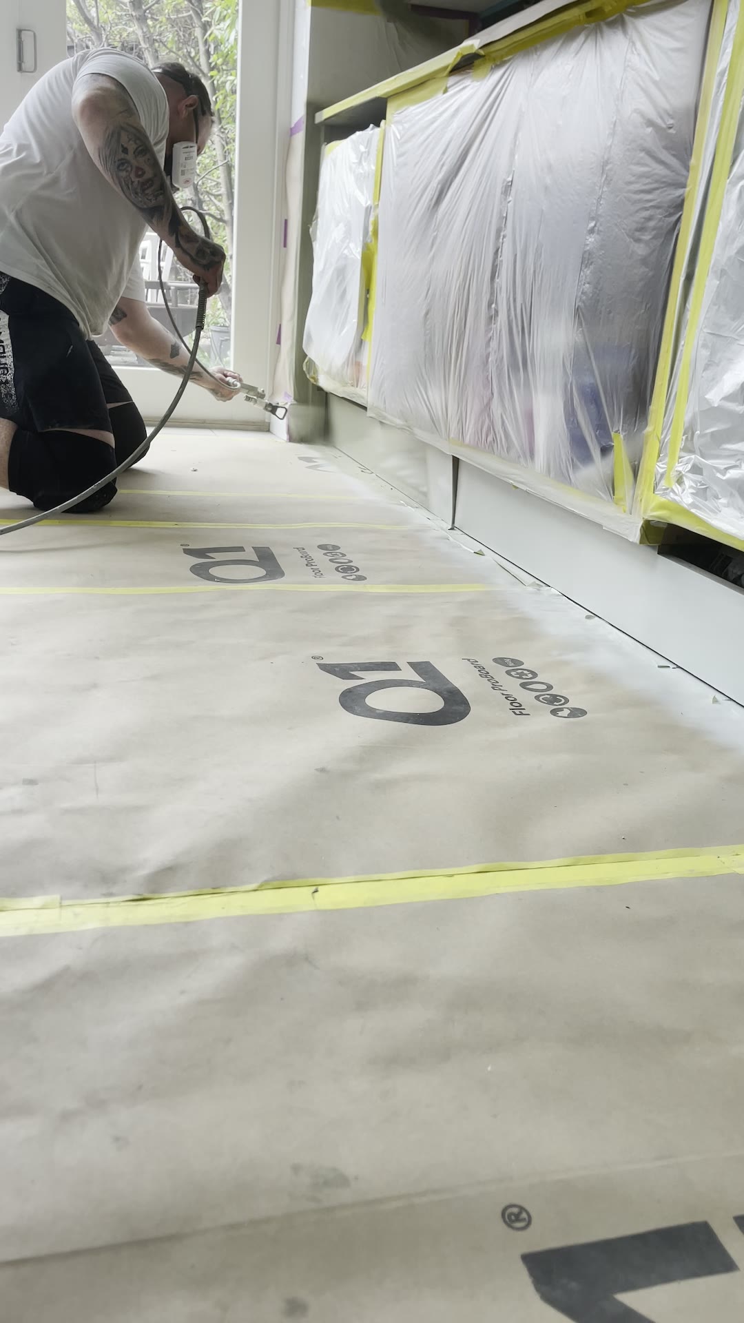 Kitchen sprayer kneeling on protective floor board spraying a run of base unit plinths and end panels